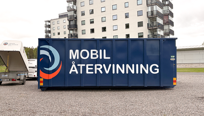 En bild på den mobila återvinningscentralen, ståender på ett grusunderlag vid ett besök ute i kommunen. I bakgrunden syns lägenhetshus.
